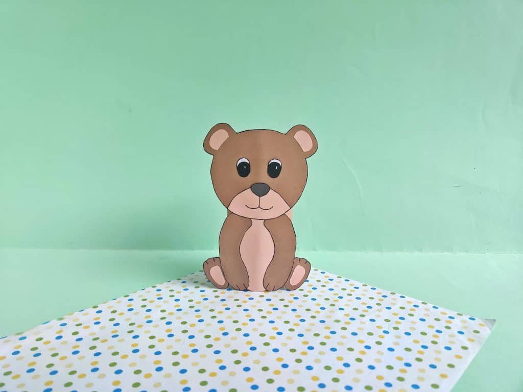 bear made from toilet paper roll and brown construction paper on a green background and a polka dotted sheet of paper below it