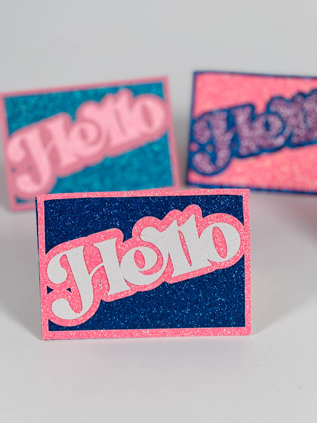 five glitter greeting cards in various color combos, all saying “Hello” in bold script font.