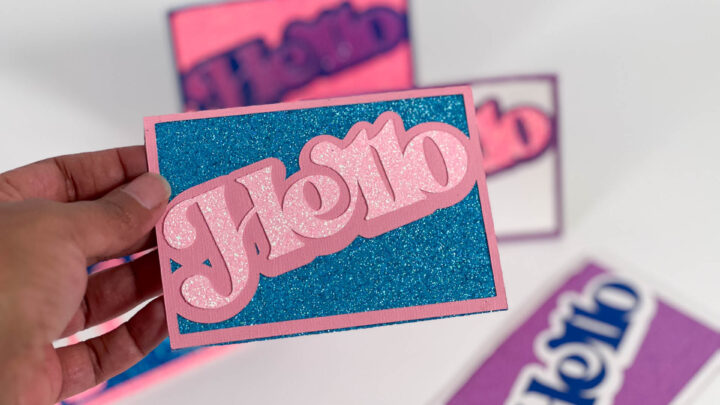 Close-up of a handmade pink and blue glitter &ldquo;Hello&rdquo; card being held, showing layers and texture.