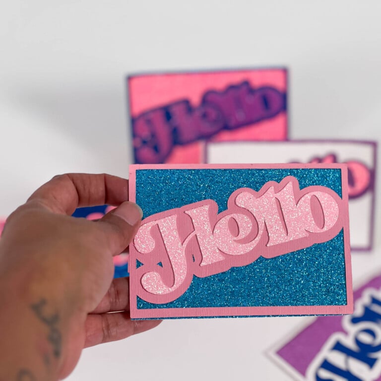 Handmade Cricut cards lined up in background with close focus on a blue and pink glitter “Hello” design.