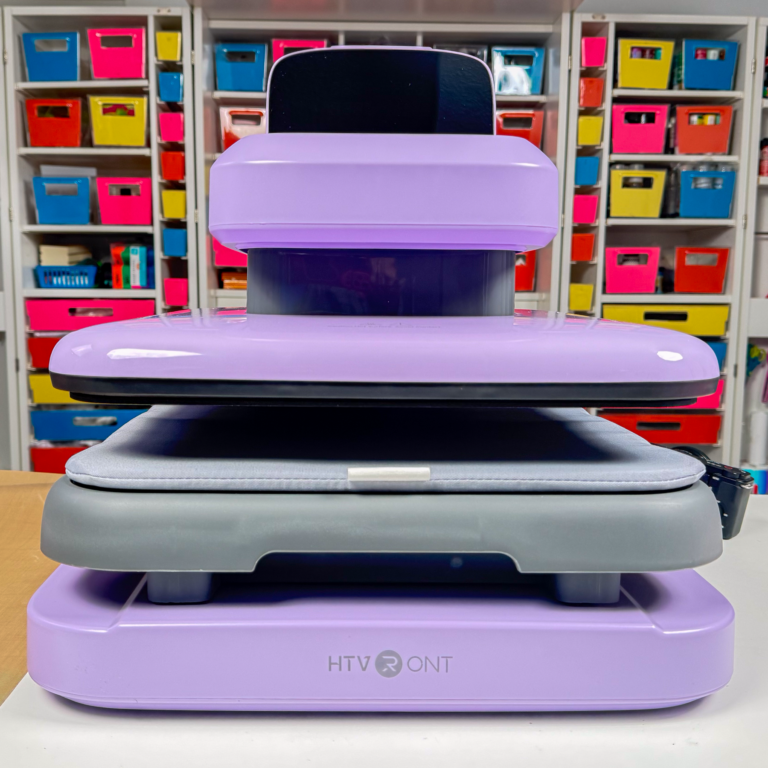 HTVRONT Auto Heat Press 2 in lavender purple, front view, drawer closed and digital display tilted up, on a craft table in front of colorful labeled storage bins